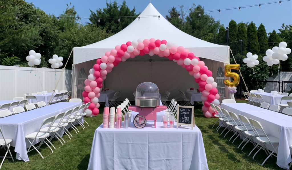 Pink cotton candy machine cart on a white linen table in front of a white party tent with a pink and white balloon arch and white folding tables set up in a Nassau County backyard for a kids birthday party